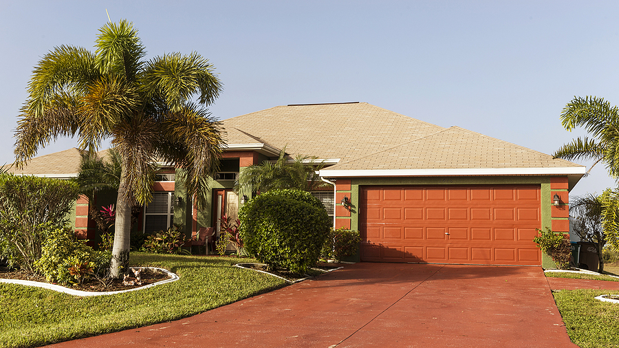 A Florida concrete block home features a red concrete driveway, a red garage door, and a beige shingle roof. It has palm trees in the front yard.